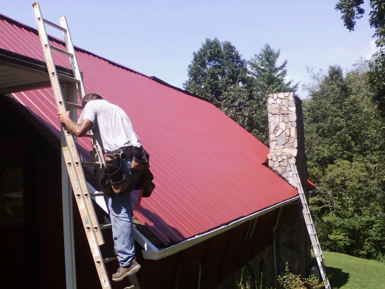 Roofer on a ladder beside a red metal roof and stone chimney — Soffit & Fascia Repair by Roofing Brothers in Jemez Pueblo